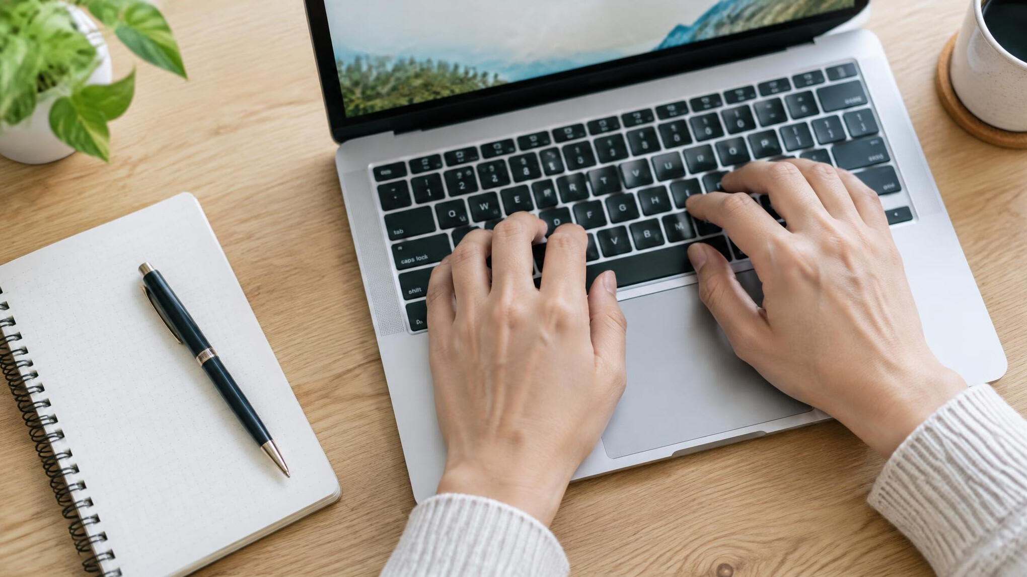Hands typing on a laptop on a wooden desk next to a notebook